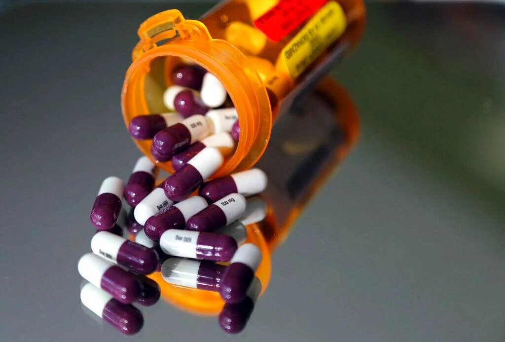 Prescription bottle spilled with purple and white capsules scattered on a reflective surface.